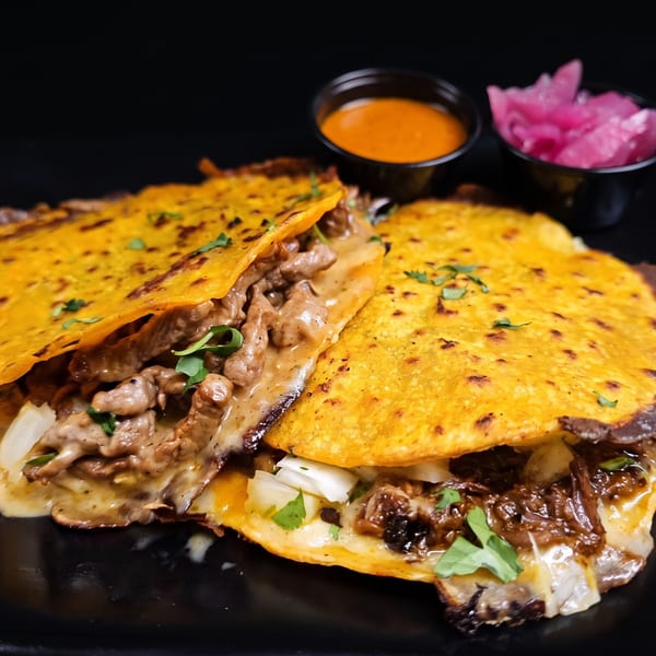 Two cheesy birria tacos with beef, onions, cilantro, and a side of dipping sauce and pickled onions.
