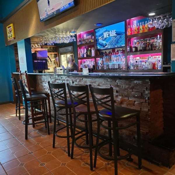 A restaurant bar with several stools, a bartender, and shelves of liquor bottles with pink lighting.