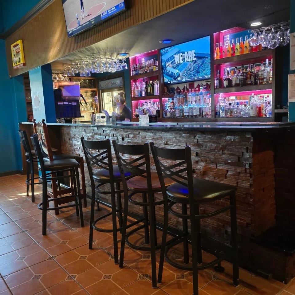A restaurant bar with several stools, a bartender, and shelves of liquor bottles with pink lighting.