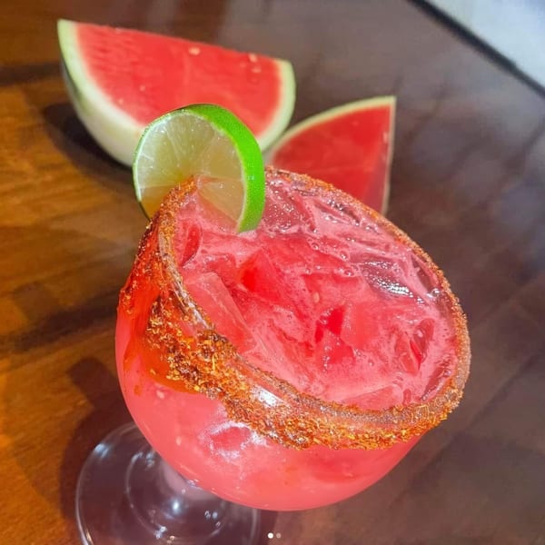 A vibrant red watermelon margarita with a chili-lime rim and a lime wedge, next to watermelon slices.