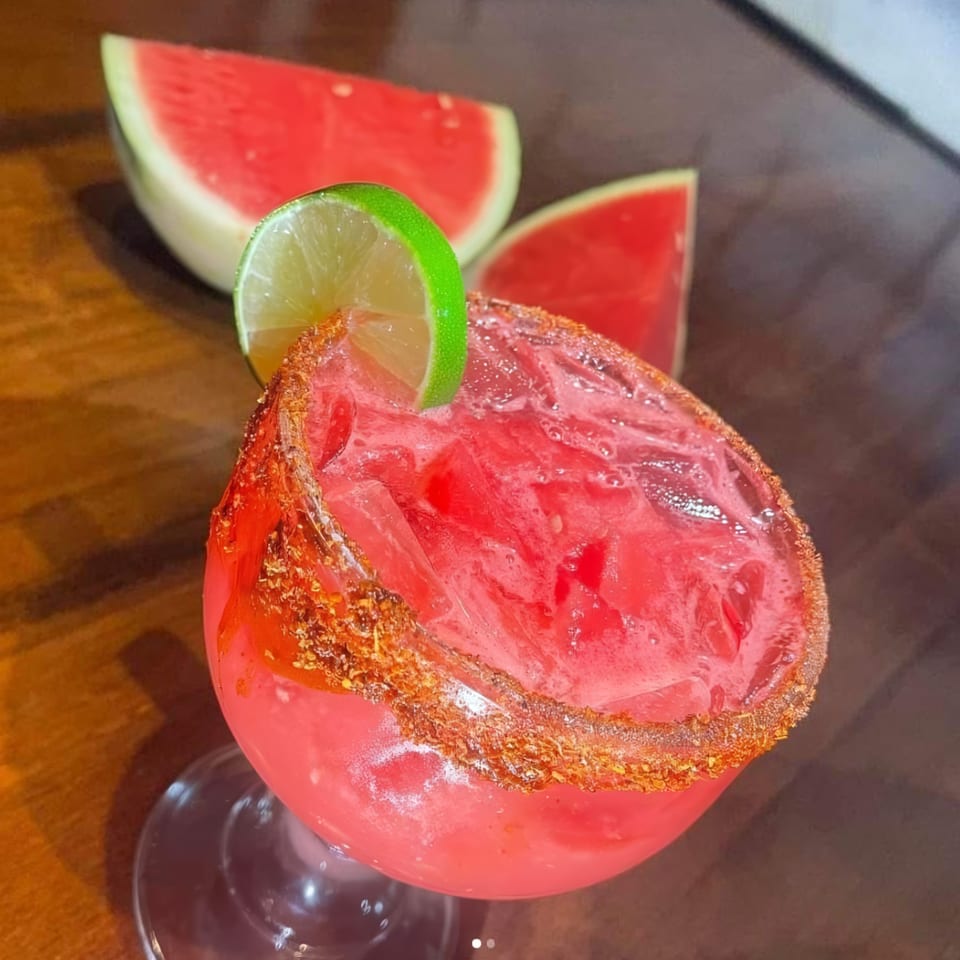 A vibrant red watermelon margarita with a chili-lime rim and a lime wedge, next to watermelon slices.
