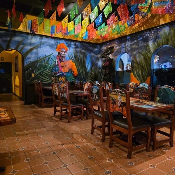 A vibrant Mexican restaurant interior with colorful papel picado, murals, and ornate wooden tables and chairs.