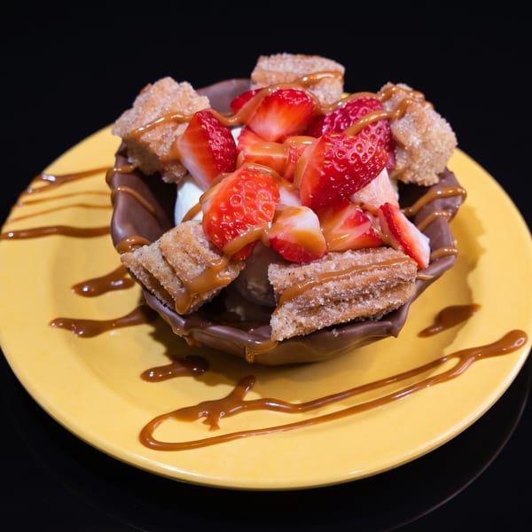 A chocolate bowl filled with ice cream, churros, strawberries, and caramel drizzle on a yellow plate.