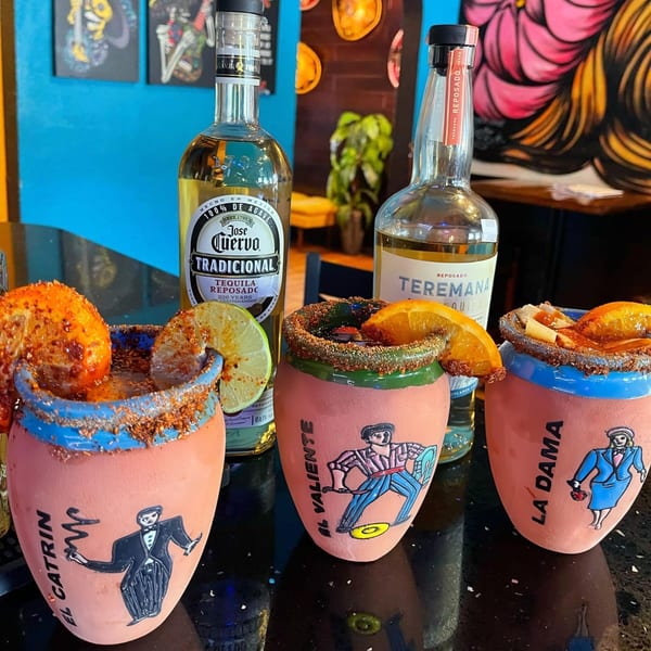 Three vibrant, decorated mugs with drinks and two tequila bottles on a dark table.
