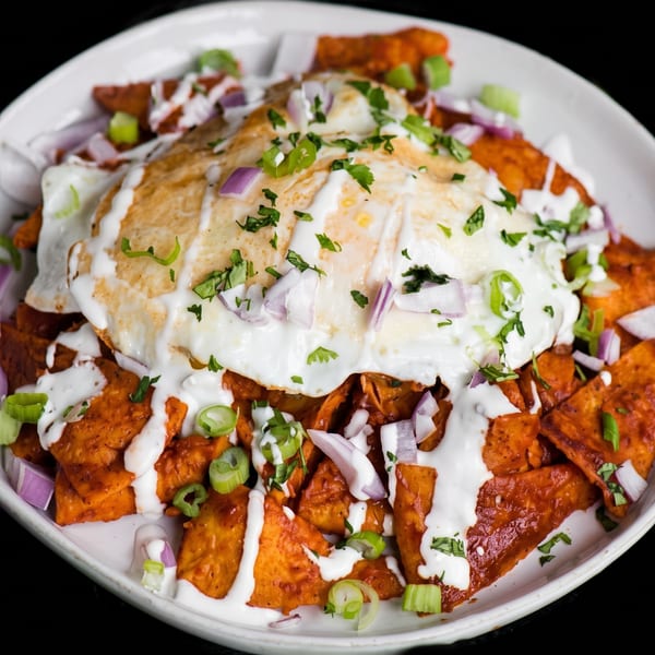 Loaded plate of spiced sweet potato chunks topped with fried eggs, creamy sauce, red onion, scallions, and herbs.