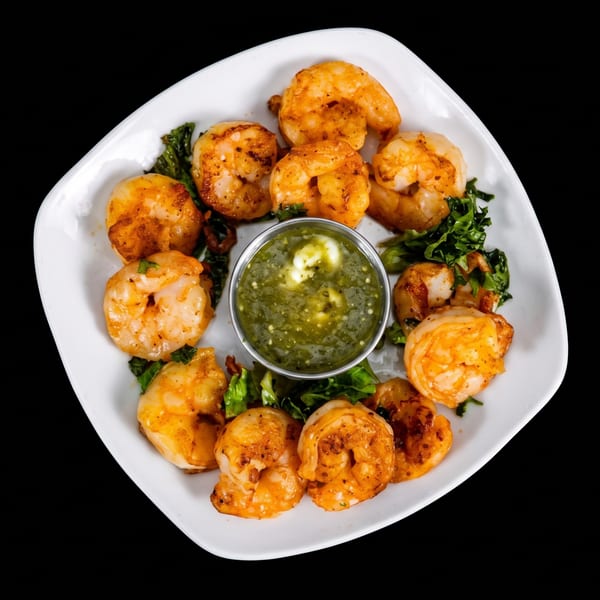 Grilled shrimp arranged around a bowl of green dipping sauce on a white plate with leafy greens.