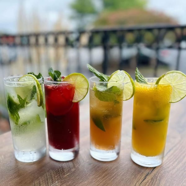 Four colorful mojitos, garnished with lime and mint, sit on a wooden table outdoors.