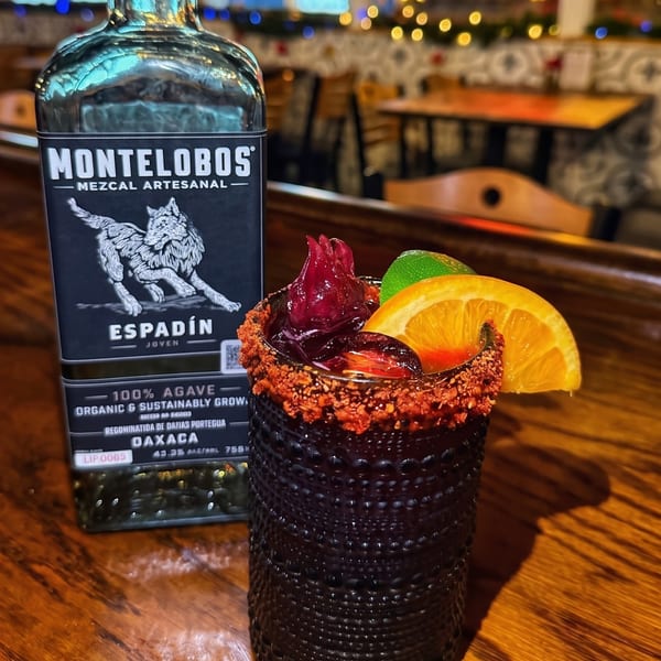 Montelobos Mezcal bottle next to a garnished cocktail with a chili-lime rim on a wooden bar.